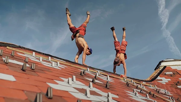 two roofers, topless, doing handstand on a roof