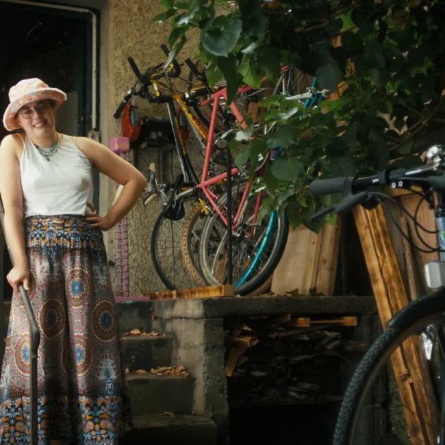 A cinematic character portrait from a Holy Moly Films production, showing actress Ellen Hascher posing on exterior steps surrounded by vintage bicycles.