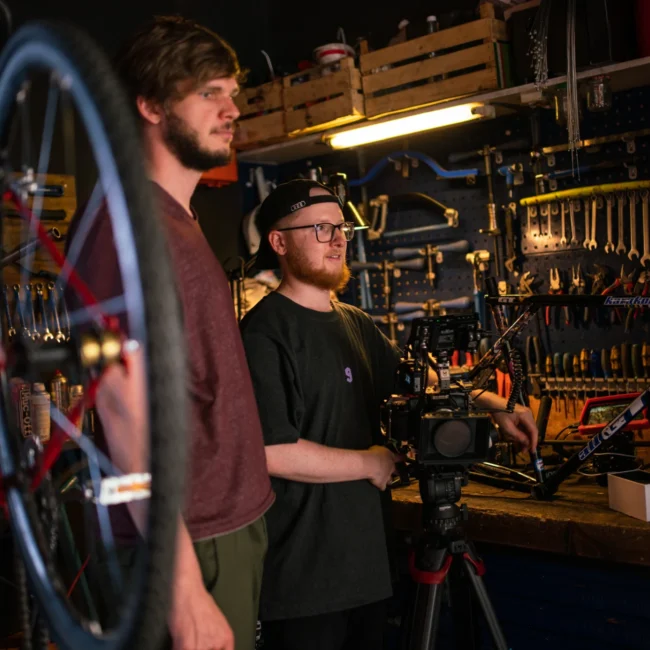 The Holy Moly Films production team, including a cinematographer with a camera rig and gaffer, planning a scene in the bike workshop.
