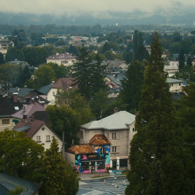 An elevated, atmospheric wide shot establishing the Salzburg neighborhood and the colorful 'Fanzy' bicycle shop nestled among trees and houses.