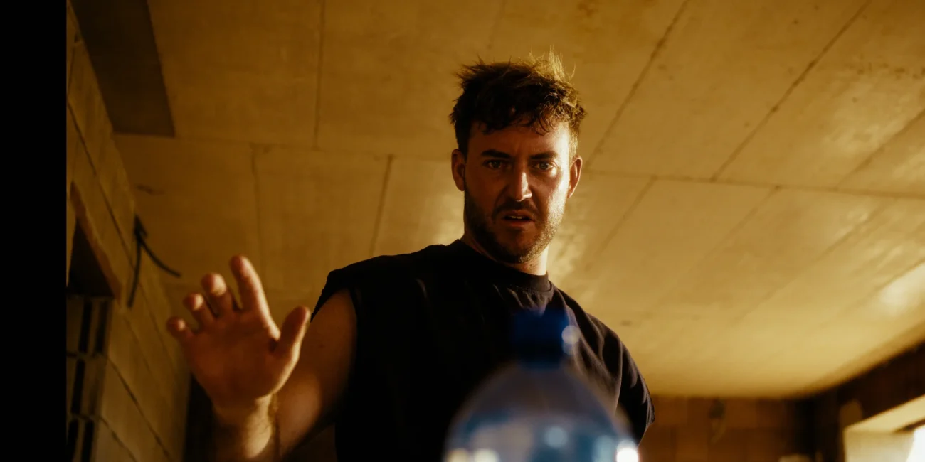 Dramatic close-up of a construction worker hesitantly reaching out toward a mystery water bottle in the Austrian Kiwi comedy sketch.
