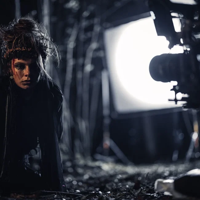 Actress in dark fantasy witch attire framed by a cinema camera with high-contrast backlight on location.