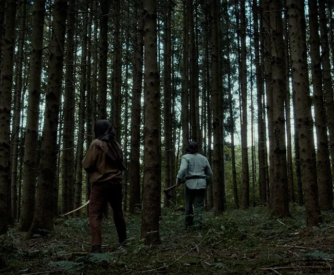 Wide cinematic shot of masked hunters equipped with a bow and axe stalking through a dense pine forest in HECATE.