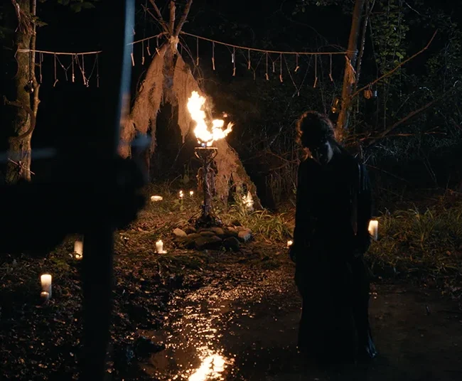 Dramatic night scene featuring a witch standing near a flaming altar with an archer aiming from the foreground shadows.