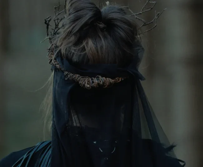 Over-the-shoulder shot of the witch's detailed headpiece, featuring a twig crown and black veil in the forest.