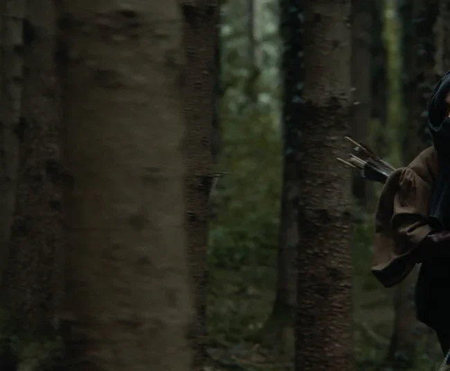 Action cinematic still of Julia Rajsp playing the masked hunter running through the woods holding a longbow.