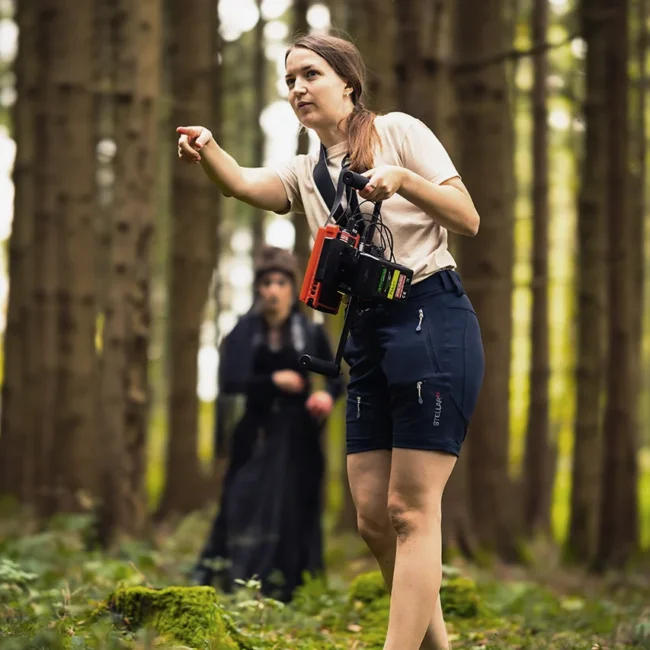Director Mascha Deikova pointing out direction on set while holding a director monitor in the forest during the HECATE short film production.