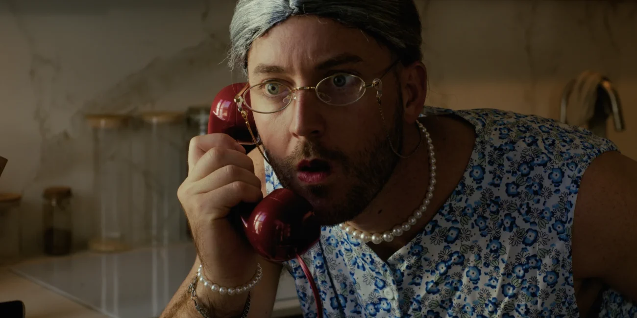 Austrian Kiwi dressed as an old woman talking shocked on a vintage red telephone in a Holy Moly Films sketch.